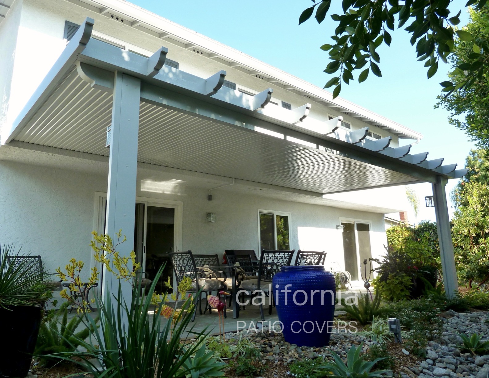 A stylish patio cover with a modern design extends over outdoor seating, surrounded by lush greenery and decorative pots, perfect for relaxation.