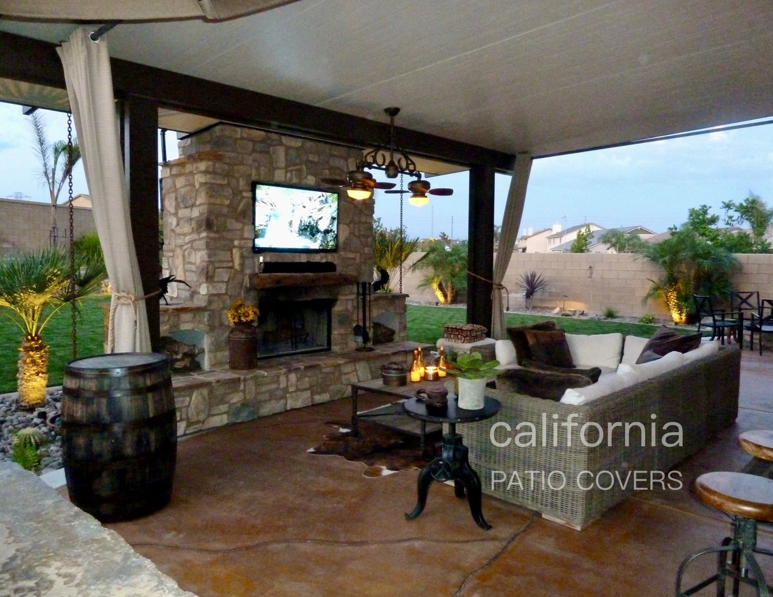 Cozy outdoor living space with a stone fireplace, mounted TV, comfortable seating, and tropical plants, ideal for relaxation and entertainment.