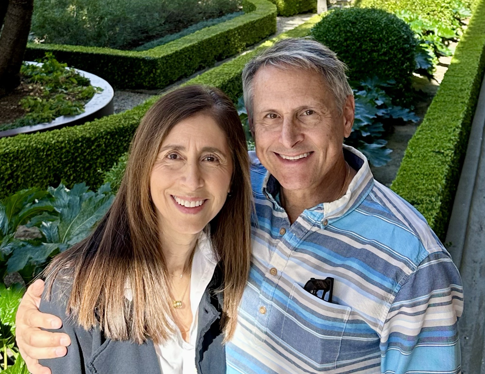 A smiling couple poses together in a lush garden, highlighting their joy and connection amidst neatly trimmed hedges and foliage.
